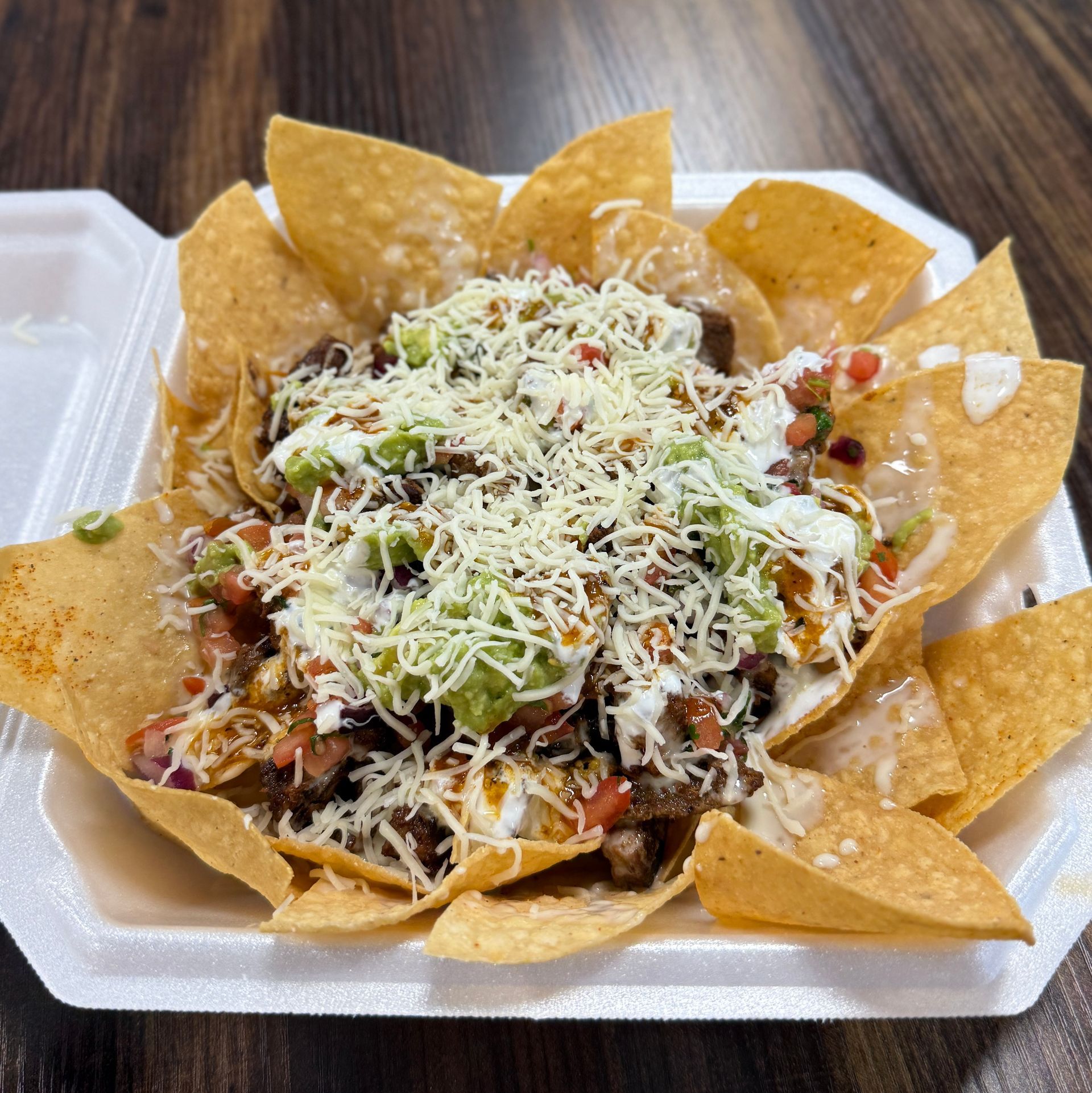 Nachos in a white container: tortilla chips topped with guacamole, pico de gallo, cheese, and sour cream.