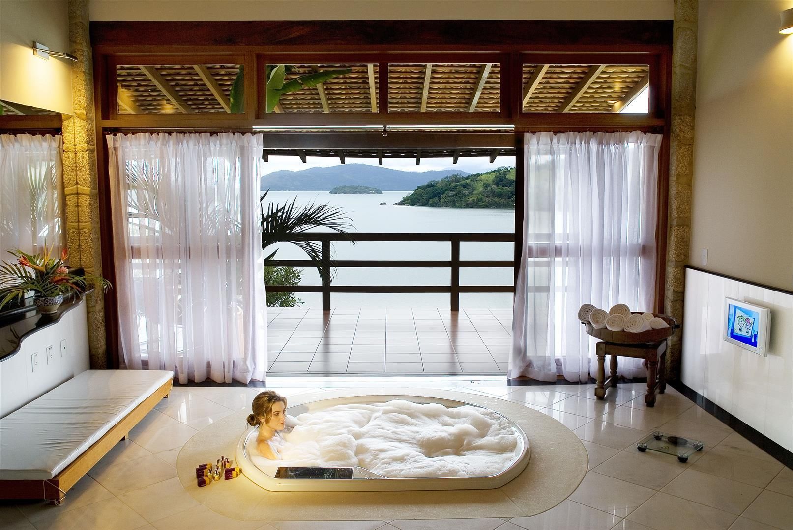 Quarto de spa com banheira redonda, vista para o mar através de cortinas abertas e TV na parede.