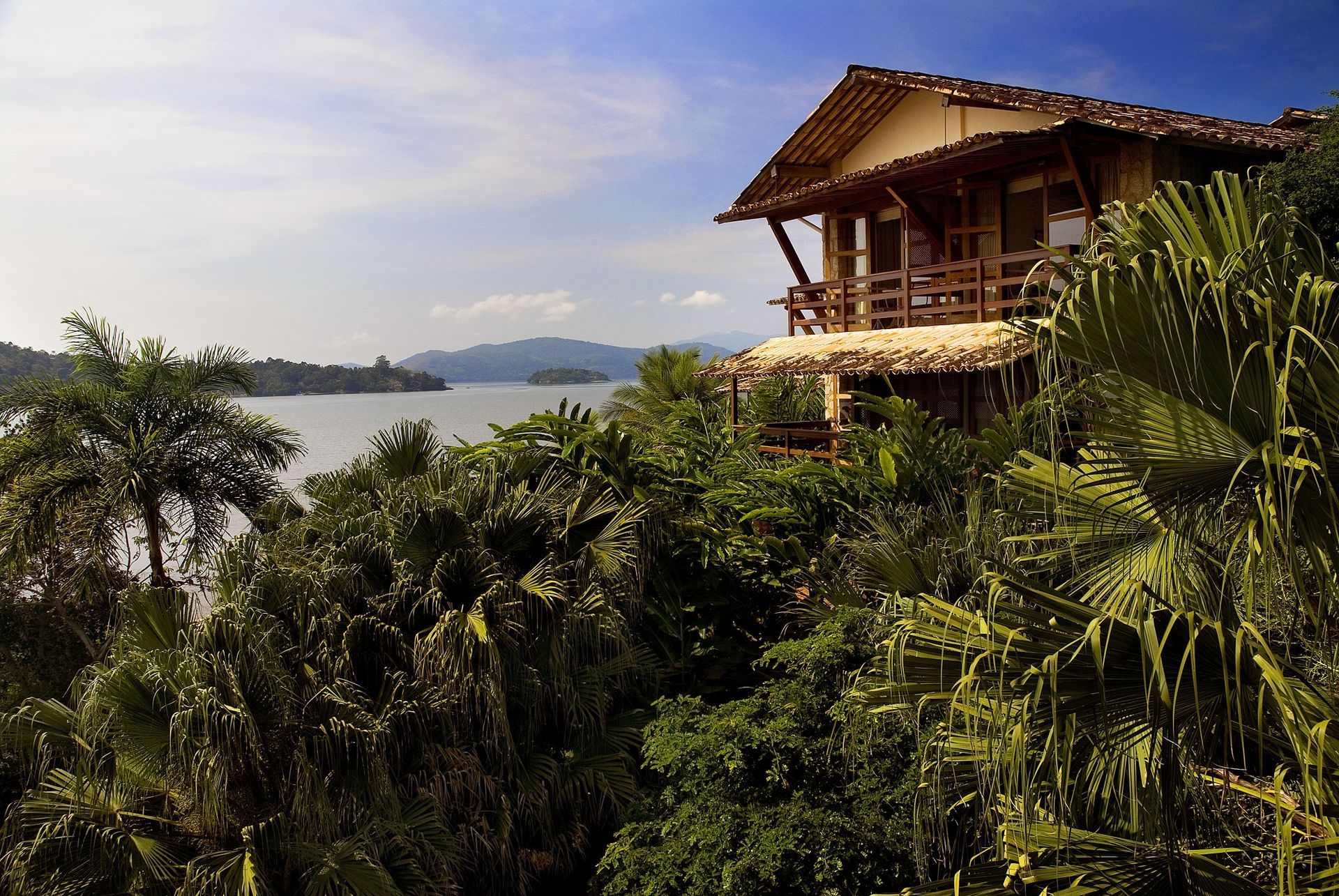 Chalé tropical na encosta com vista para uma baía tranquila, emoldurado por palmeiras e árvores verdejantes ao pôr do sol.