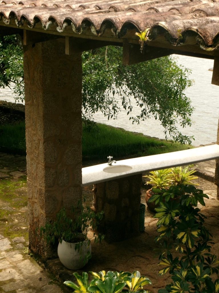 Pátio de pedra com telhado de telha, corrimão branco e vasos de plantas com vista para um jardim e lago.