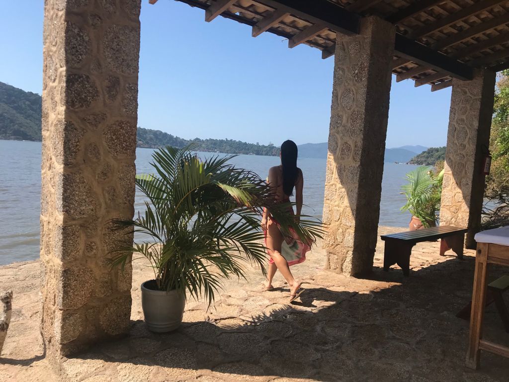 Pessoa em pé sob um alpendre sombreado com vista para uma praia ensolarada e água azul.