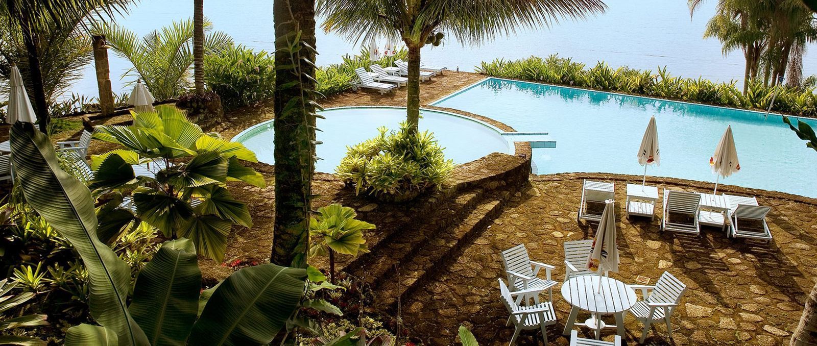 Piscina de resort tropical com palmeiras, espreguiçadeiras e um terraço com vista para o mar.