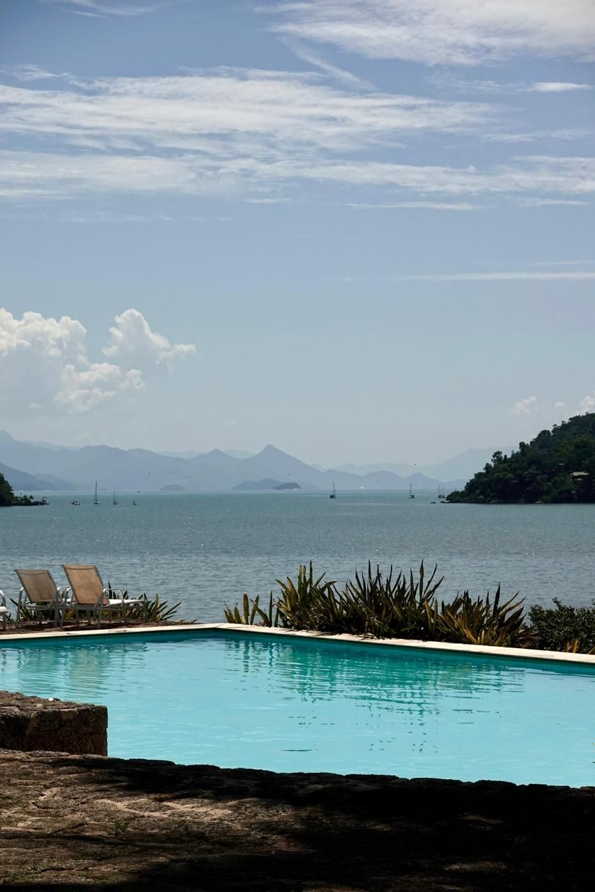 Piscina turquesa com vista para uma baía tranquila, espreguiçadeiras, ilhas ao longe e um céu límpido.