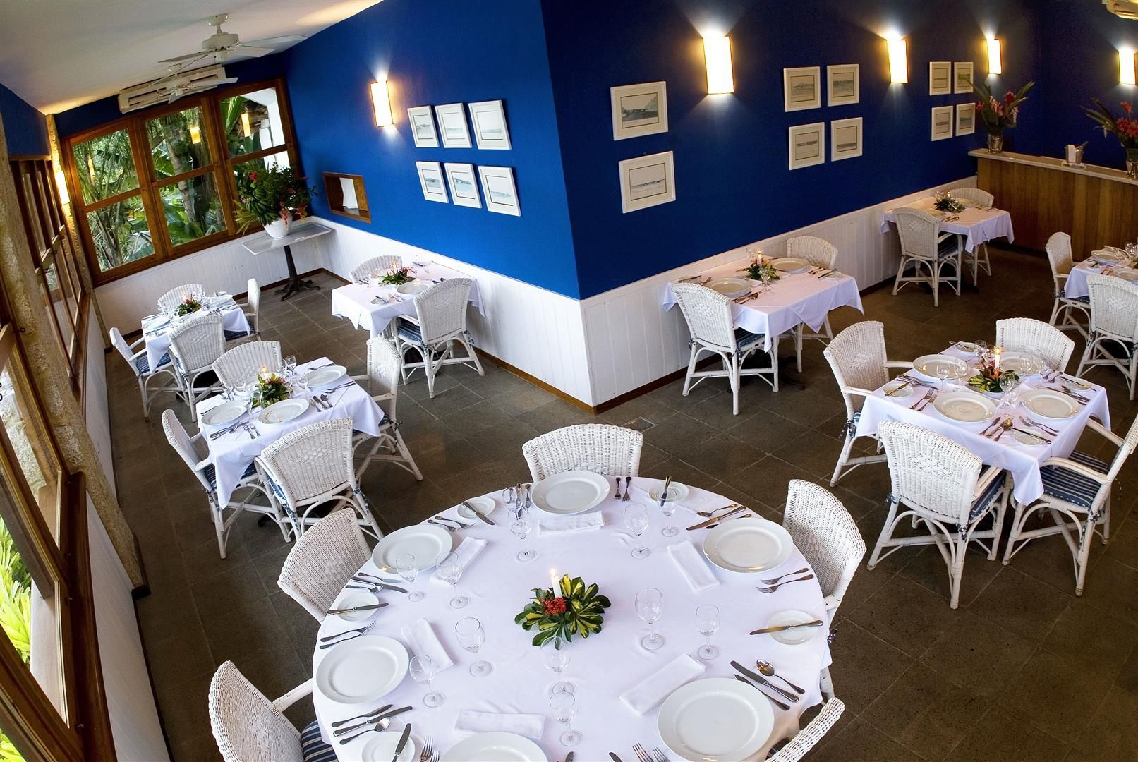 Sala de jantar bem iluminada com mesas cobertas de branco, paredes azuis e um arranjo floral central.