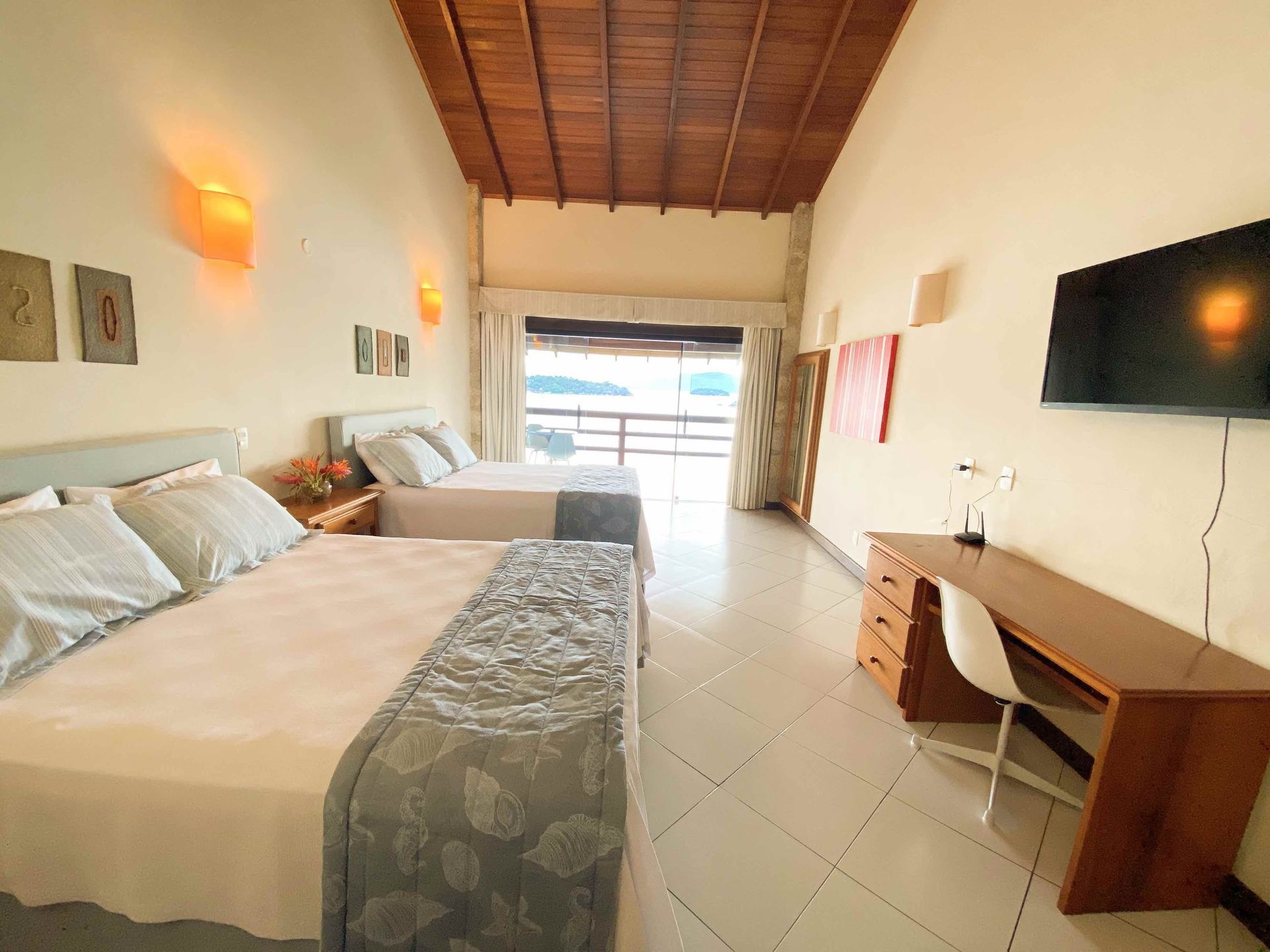 Quarto de hotel espaçoso com duas camas, uma escrivaninha, TV de parede e porta de vidro deslizante para uma varanda.