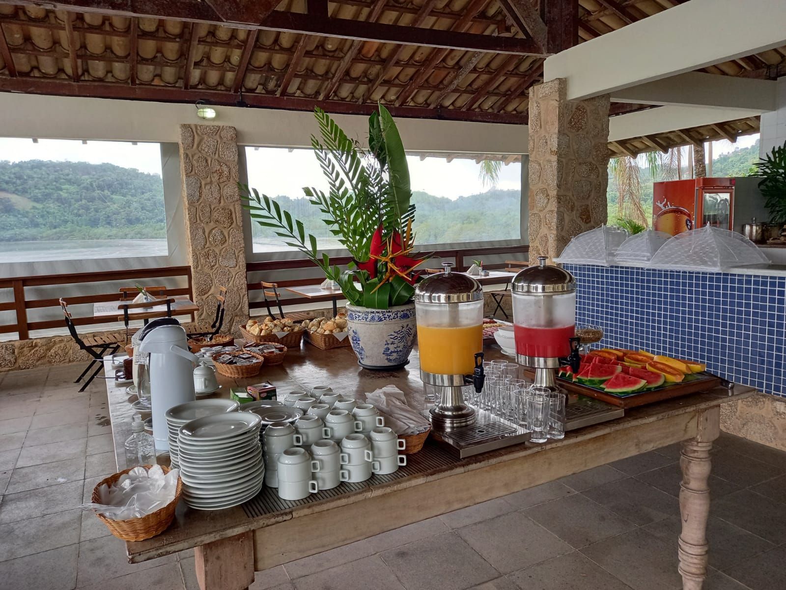 Mesa de buffet ao ar livre com xícaras de café, bebidas e petiscos com vista para um lago e colinas.