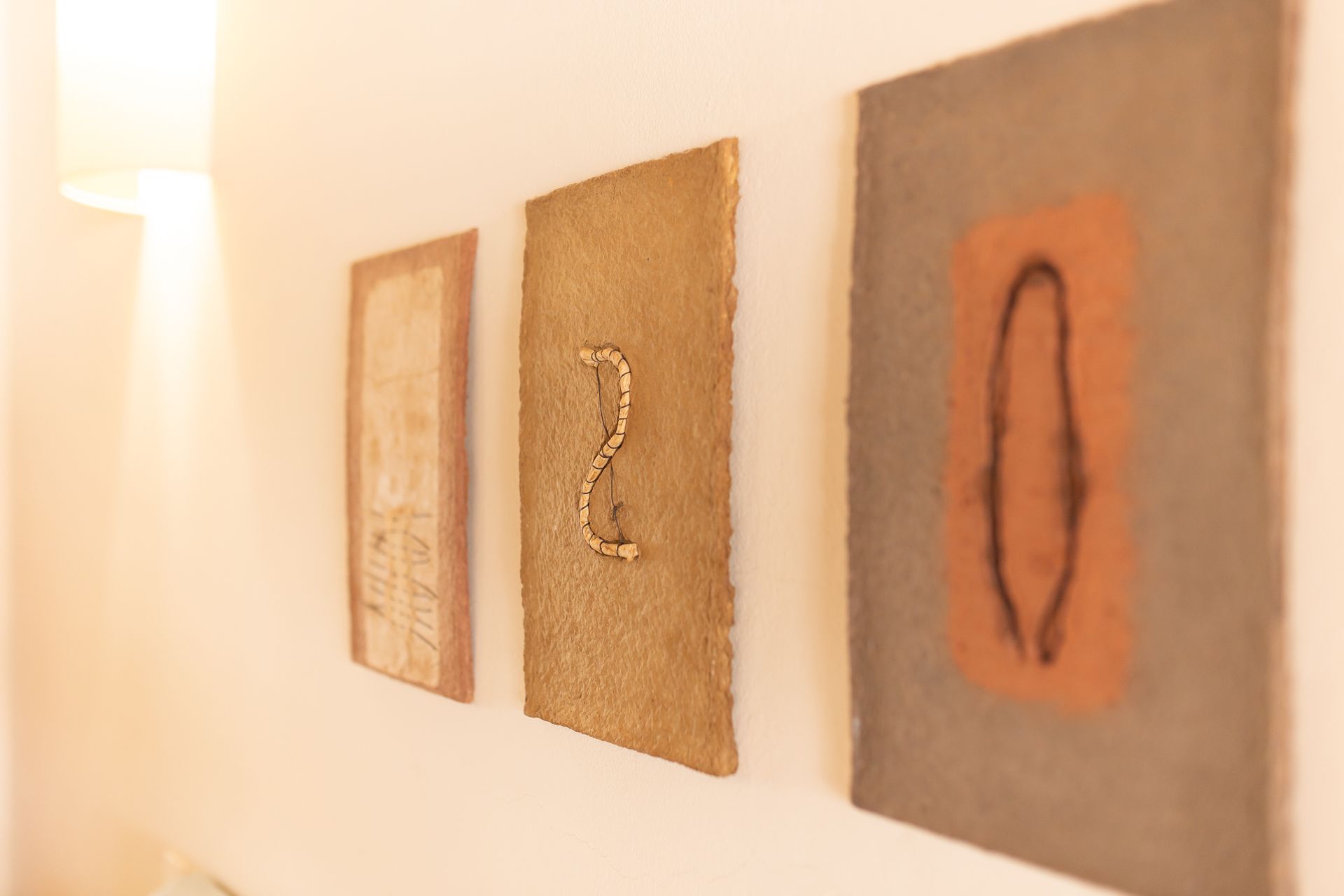 Três obras de arte emolduradas com texturas, sobre uma parede branca, iluminadas pela luz quente do sol.