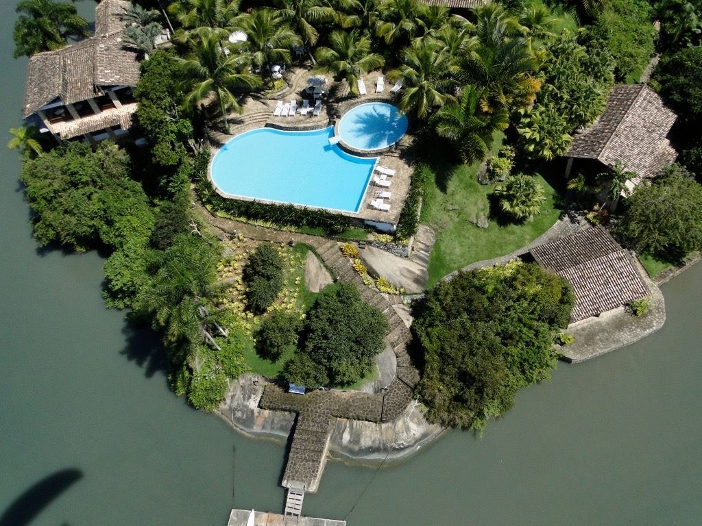 Vista aérea de um resort numa ilha tropical com uma grande piscina, palmeiras e pequenas cabanas de palha perto da água.