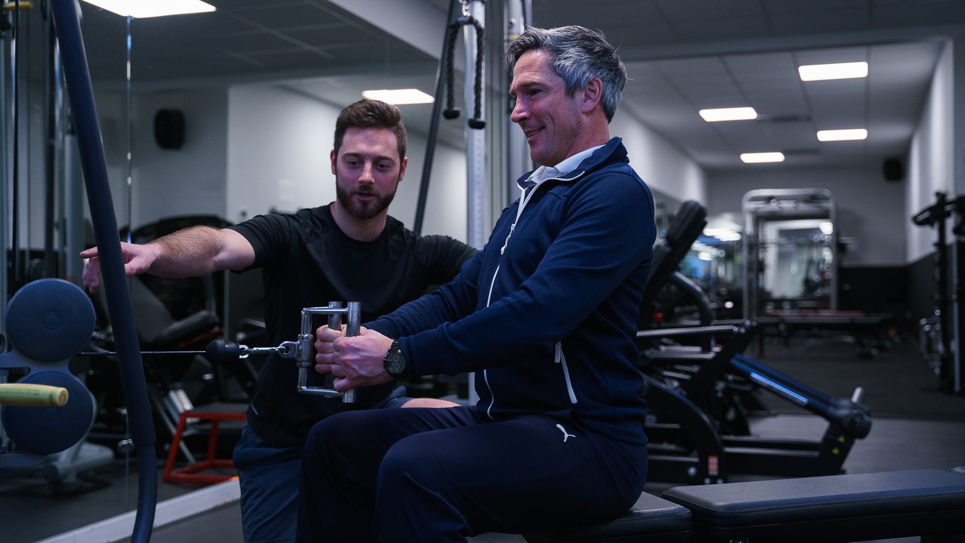 Een man zit op een fitnessapparaat in een sportschool terwijl een andere man hem helpt.