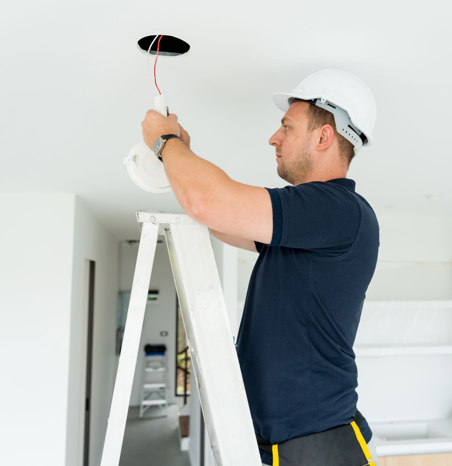 Worker in a hard hat installing a ceiling light on a ladder in a bright room