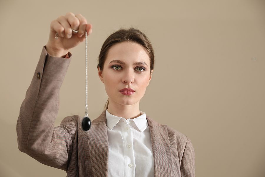 Woman in blazer holds a pendulum, gazing intently. Beige background.