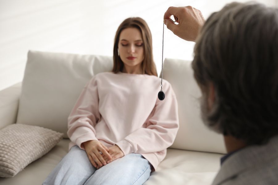 Woman undergoing hypnosis with a pendulum, eyes closed, on a white couch.