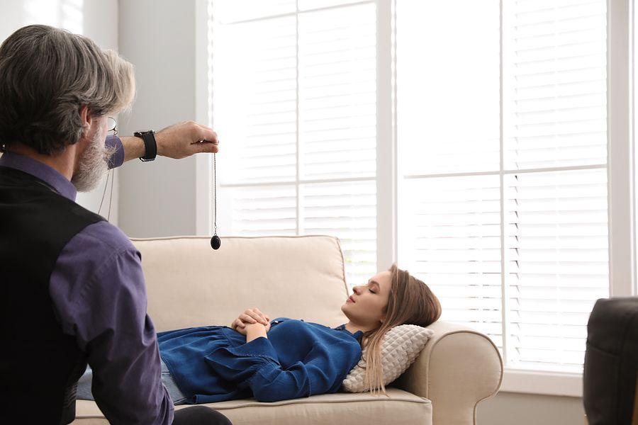 A man hypnotizes a woman lying on a couch in a room with a window.