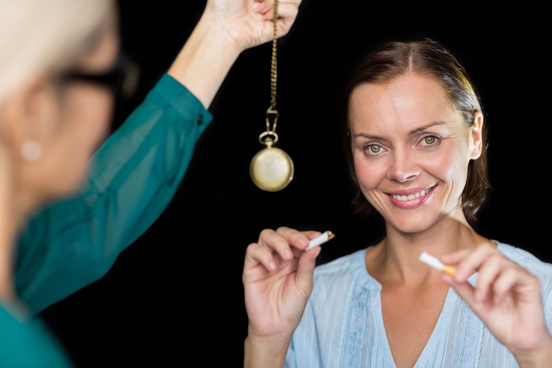 Woman smiles, breaking a cigarette, as a therapist holds a pocket watch; black background.