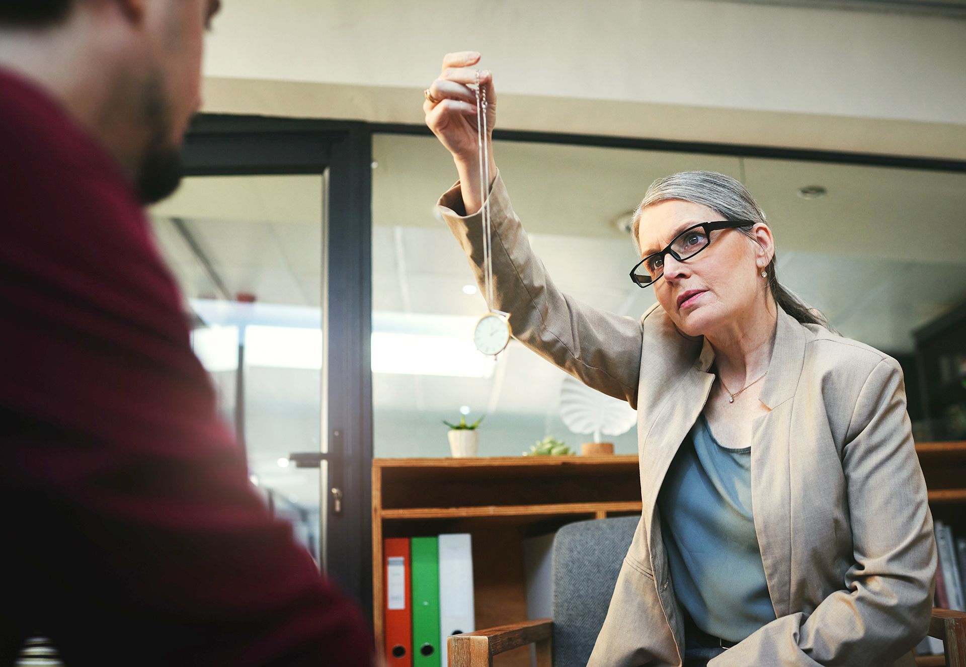 Woman with glasses and blazer uses a pendulum on a man in an office setting.