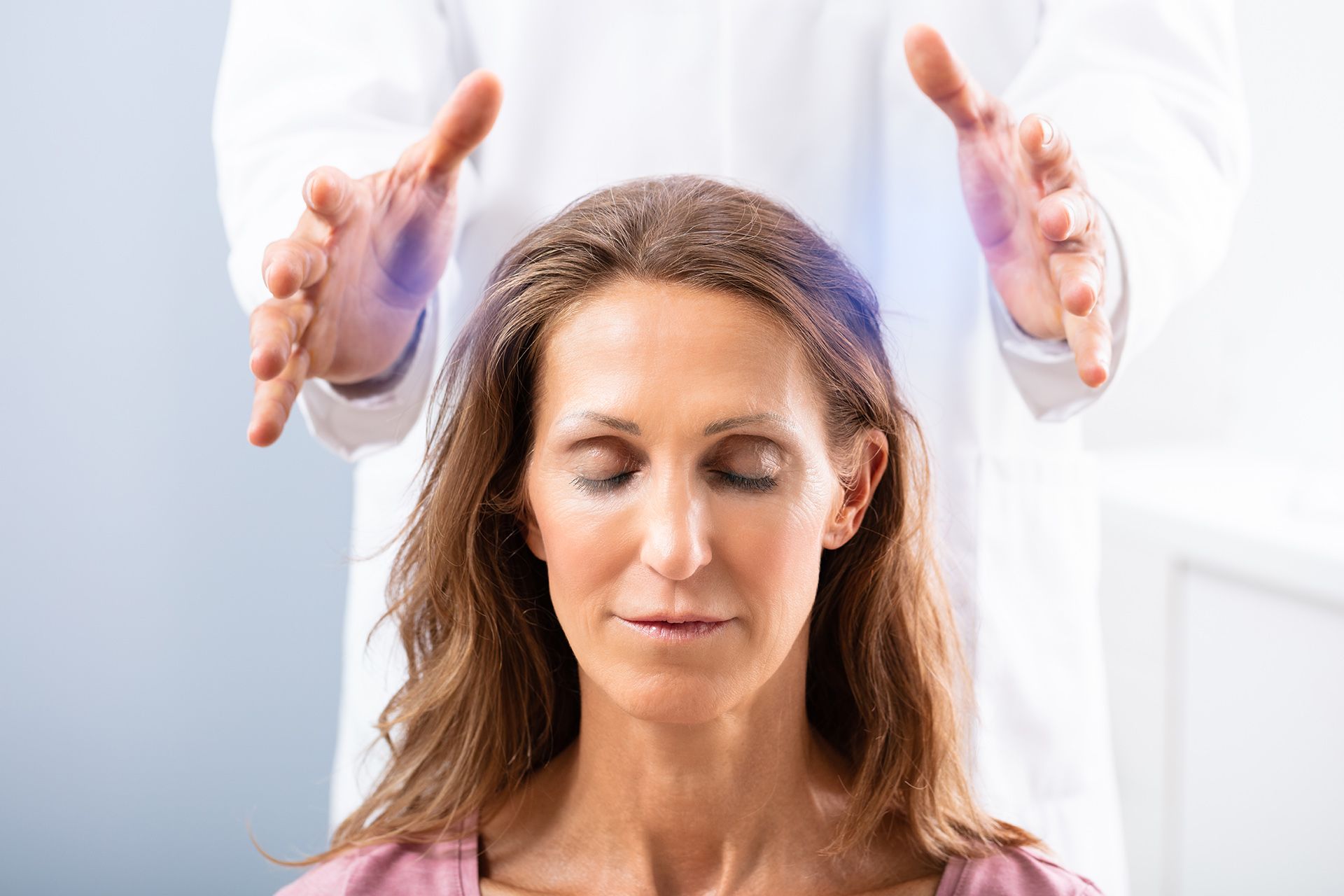 Woman receiving reiki treatment, eyes closed, with hands hovering above her head, blue light.
