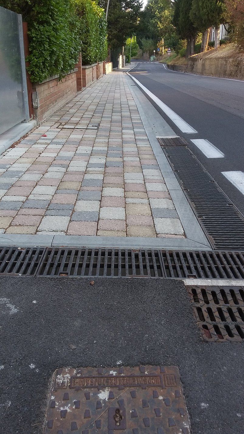 Marciapiede pavimentato con mattoni grigi, beige e marroni. Strada con linea bianca e grate. Alberi e fogliame.