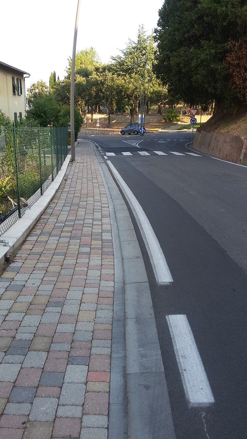 Marciapiede in mattoni accanto a una strada con attraversamento pedonale, che conduce verso alberi ed edifici.