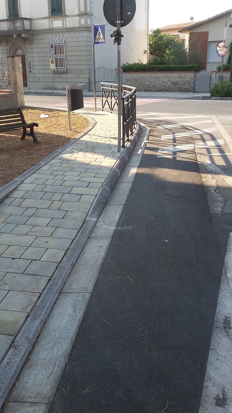 Marciapiede con superficie strutturata, un percorso in asfalto scuro e una barriera metallica vicino a una strada.