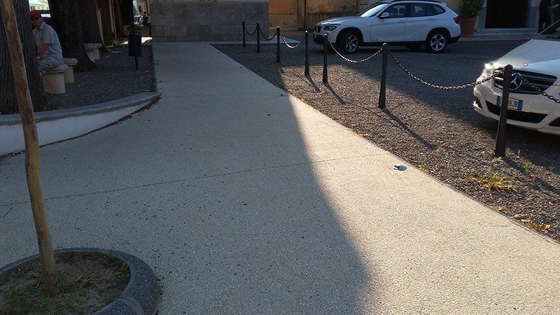 Marciapiede in cemento con paletti di segnalazione collegati da catene, due auto parcheggiate e luce solare diffusa.