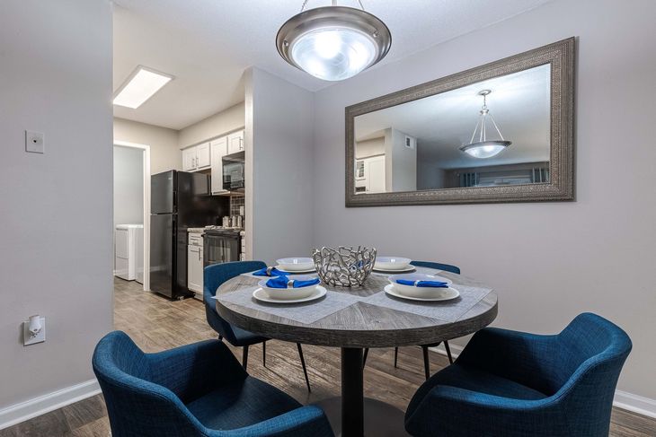Dining area with round table set for four, blue chairs, large mirror, and view of kitchen.