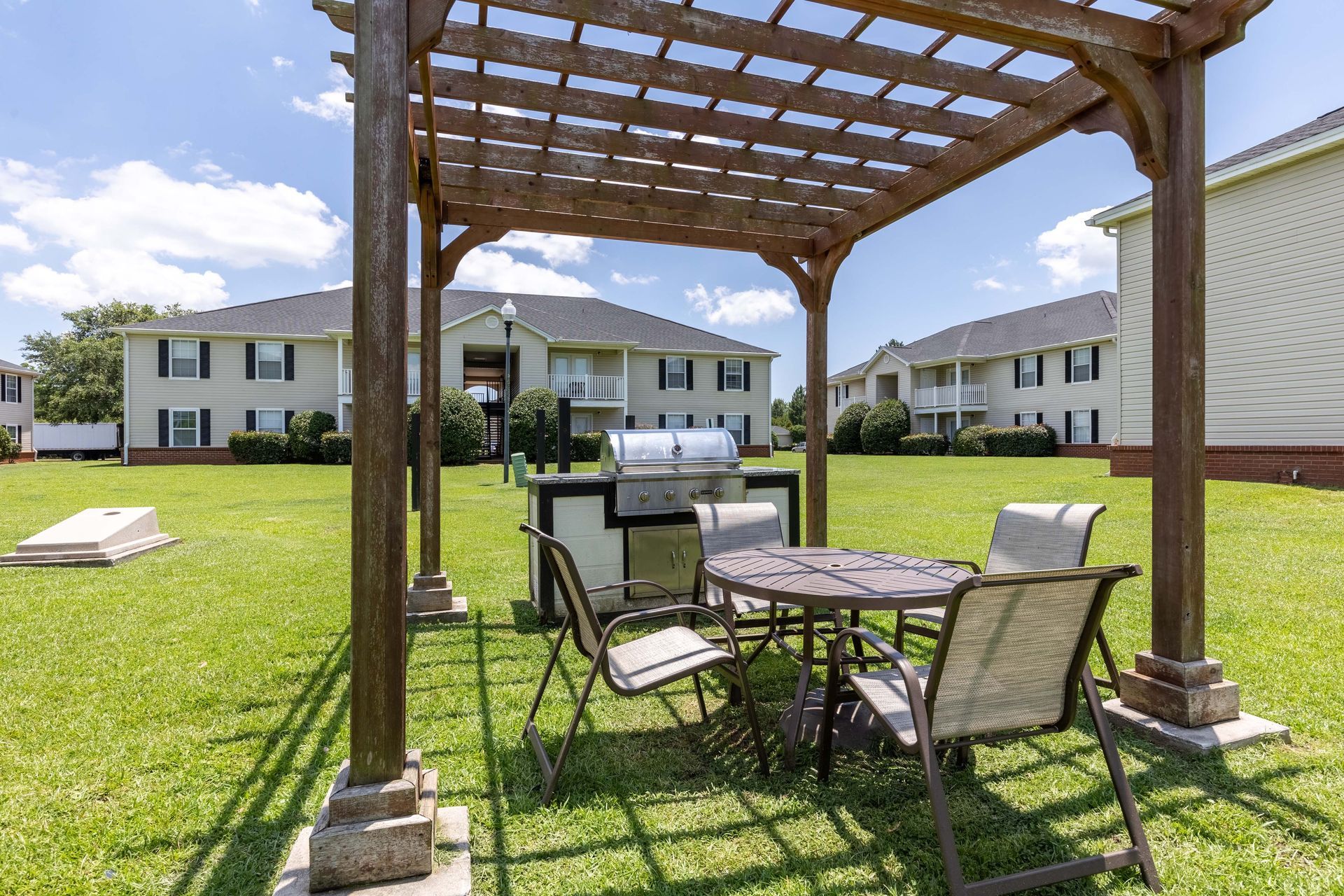 Outdoor grilling area with pergola, table, chairs, and grill, grassy area, apartment buildings in background.