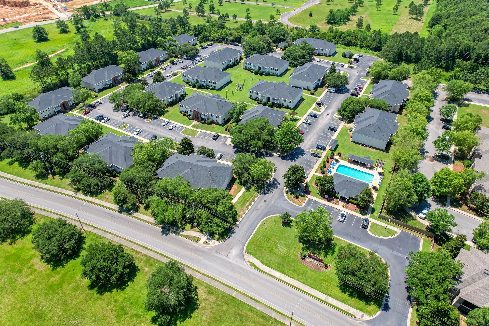 Aerial view of apartments with dark roofs, greenery, a pool, and roads.