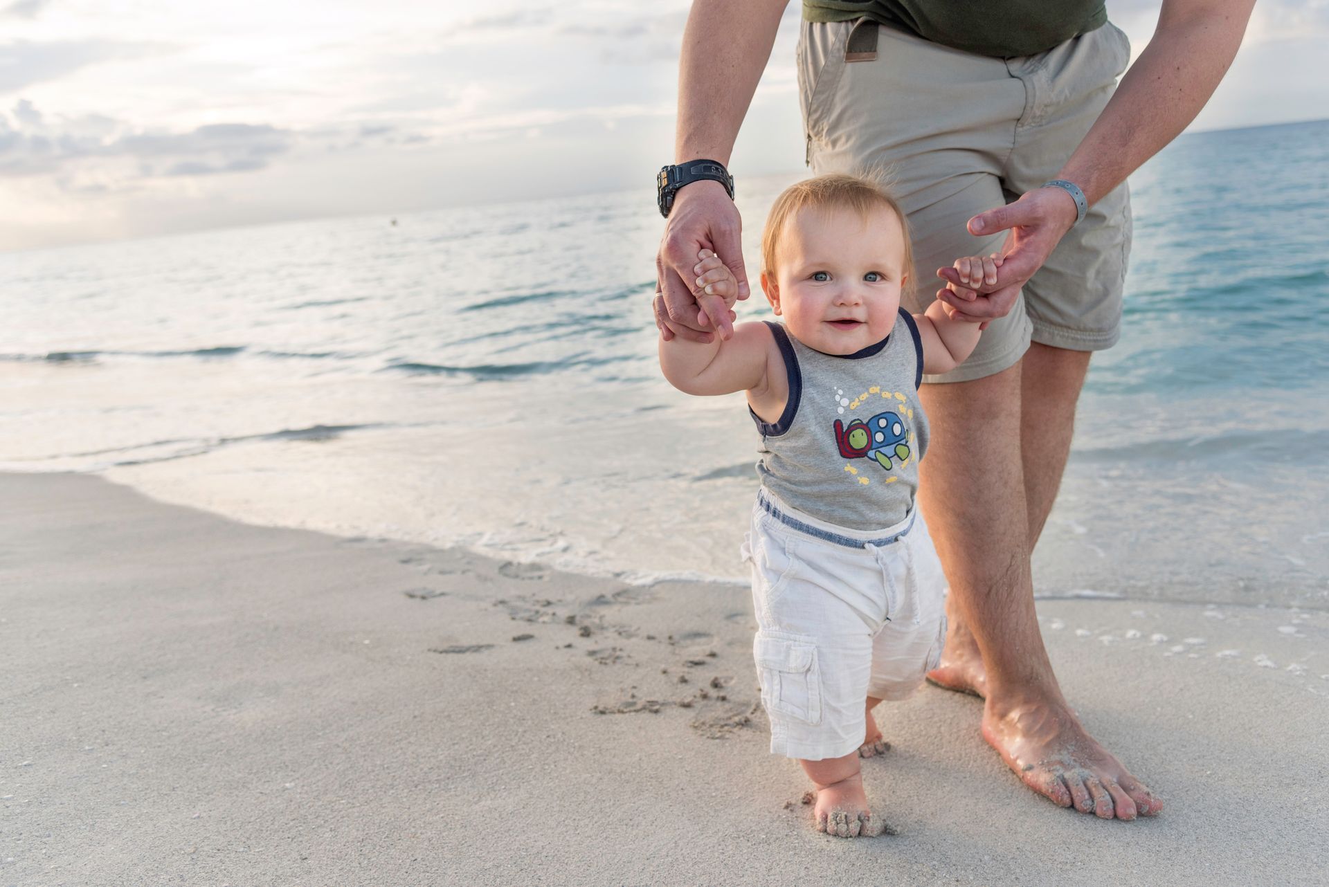 A father is walking his baby on the beach, by holding the baby's hands. Sky and water in the background.