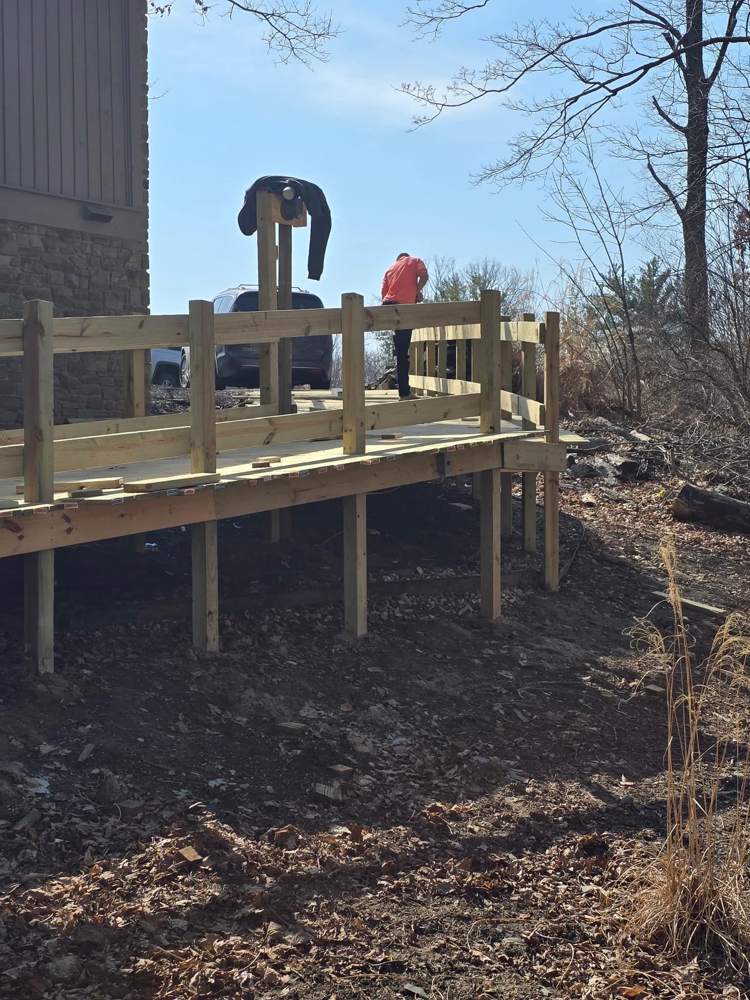 Wooden deck with railing on a hillside; person in orange near a black object under a sunny sky.