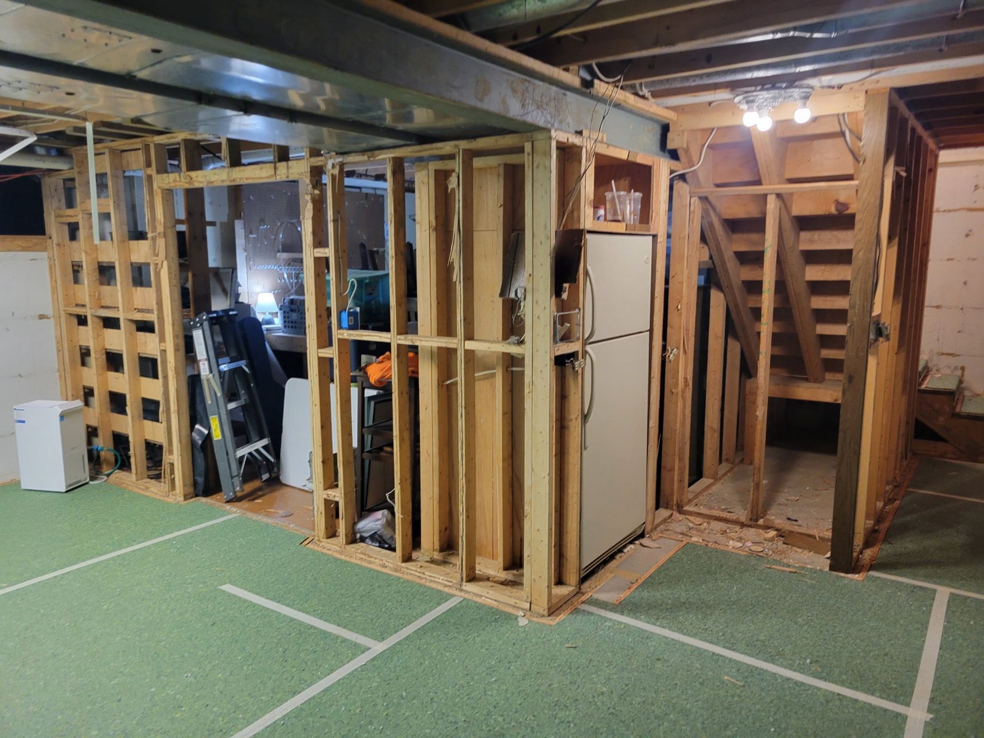 Basement renovation in progress; exposed framing, refrigerator, stairs, and green flooring.