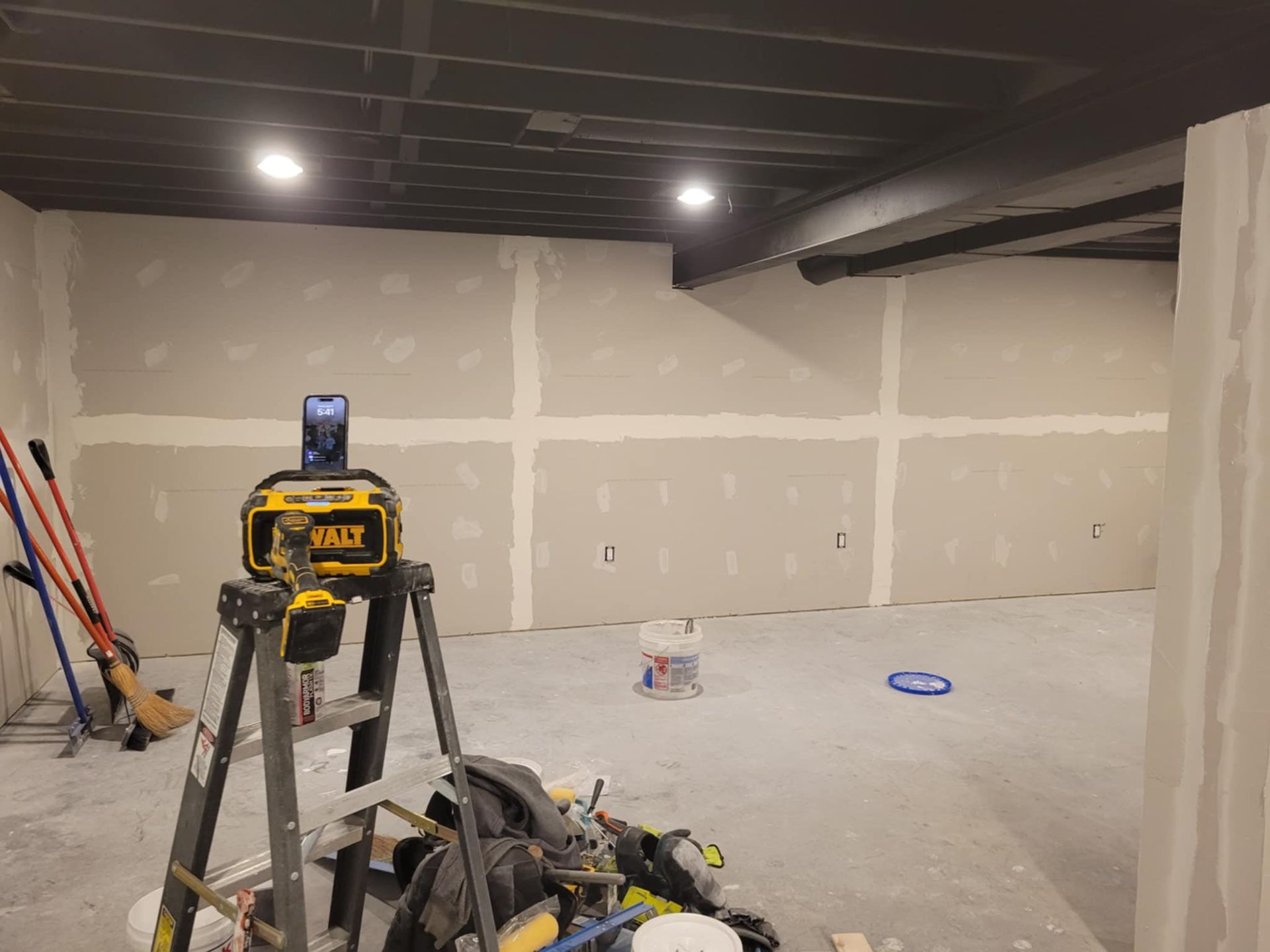 Basement under construction, drywall and tools visible. Yellow level on a ladder, black ceiling, white walls.