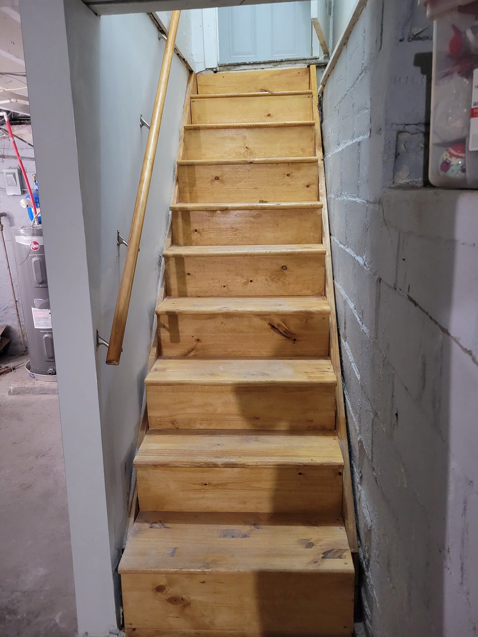 Wooden staircase leading upwards, handrail on the left. Concrete walls, unfinished basement setting.