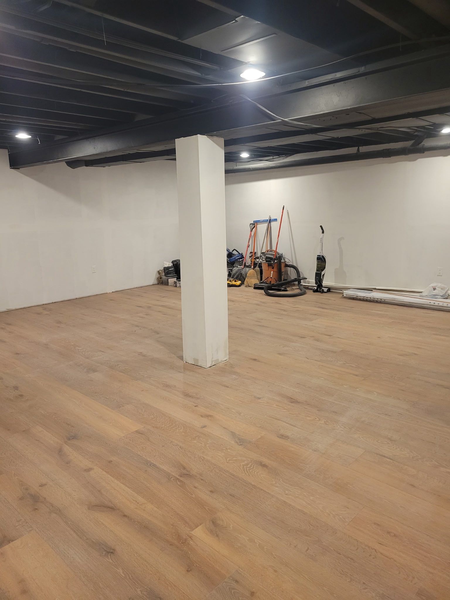 Interior view of a room under construction with white walls, wood-look floor, a central column, and tools in the background.