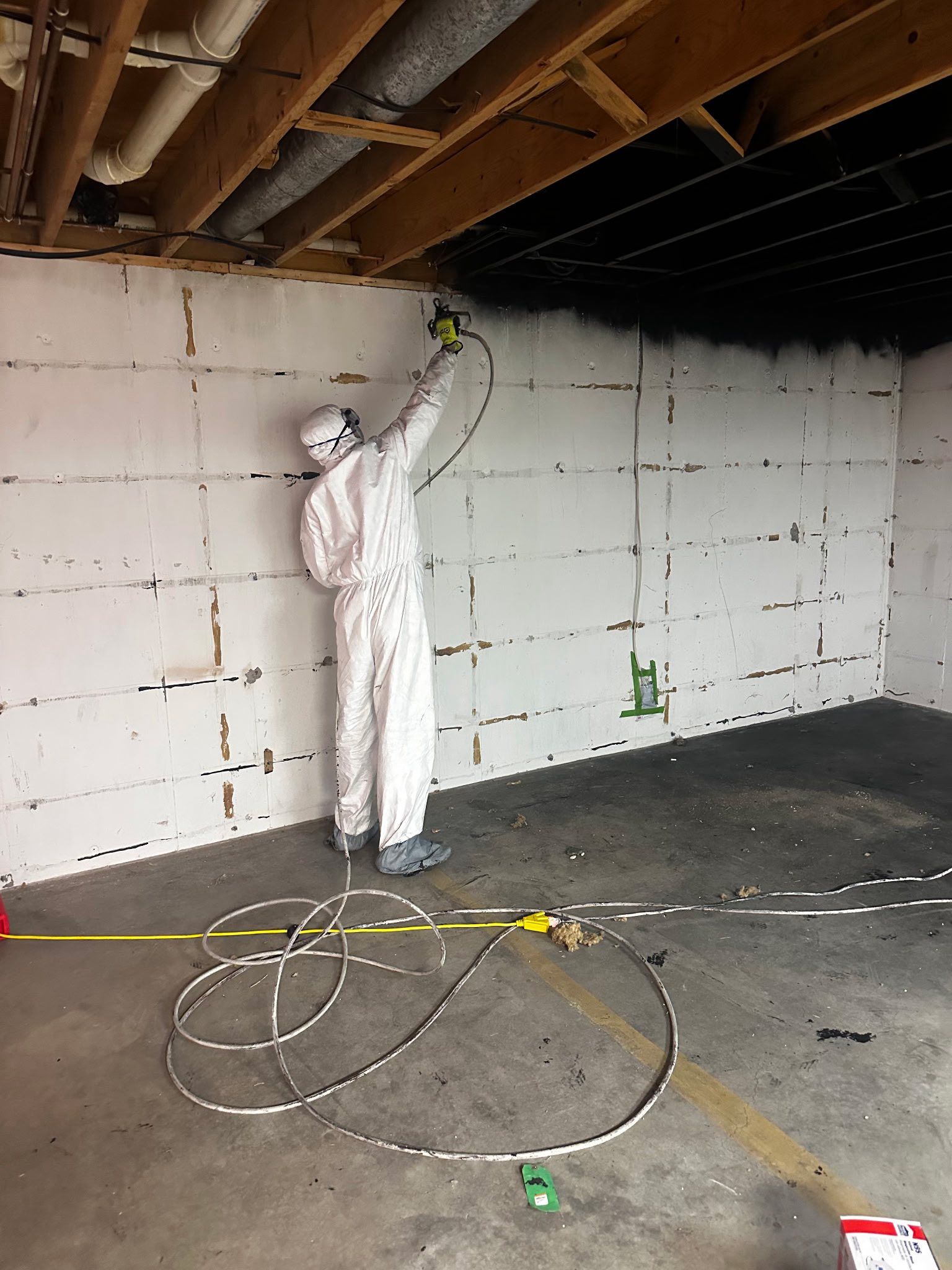 Person in protective suit spraying black material onto ceiling in a room with white foam block walls.