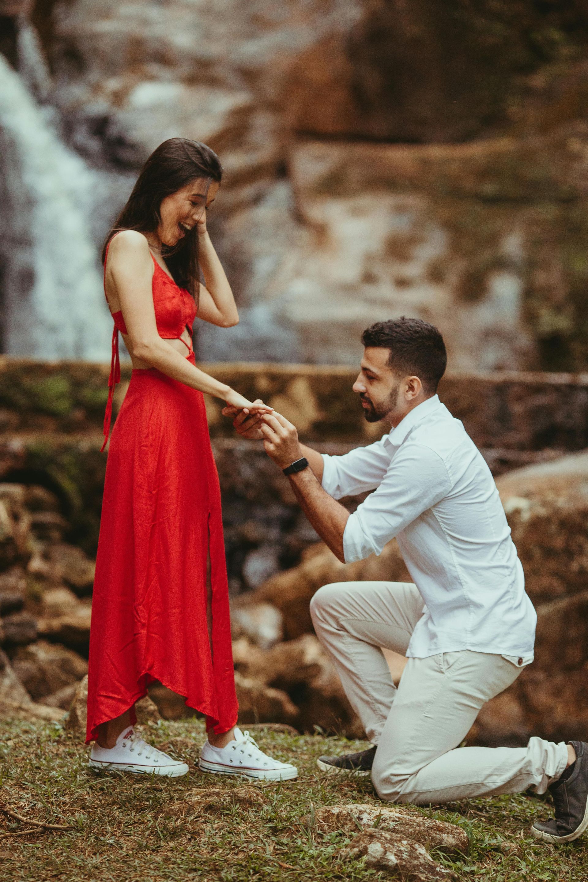 Man on one knee proposing to a woman in a red dress near a waterfall.
