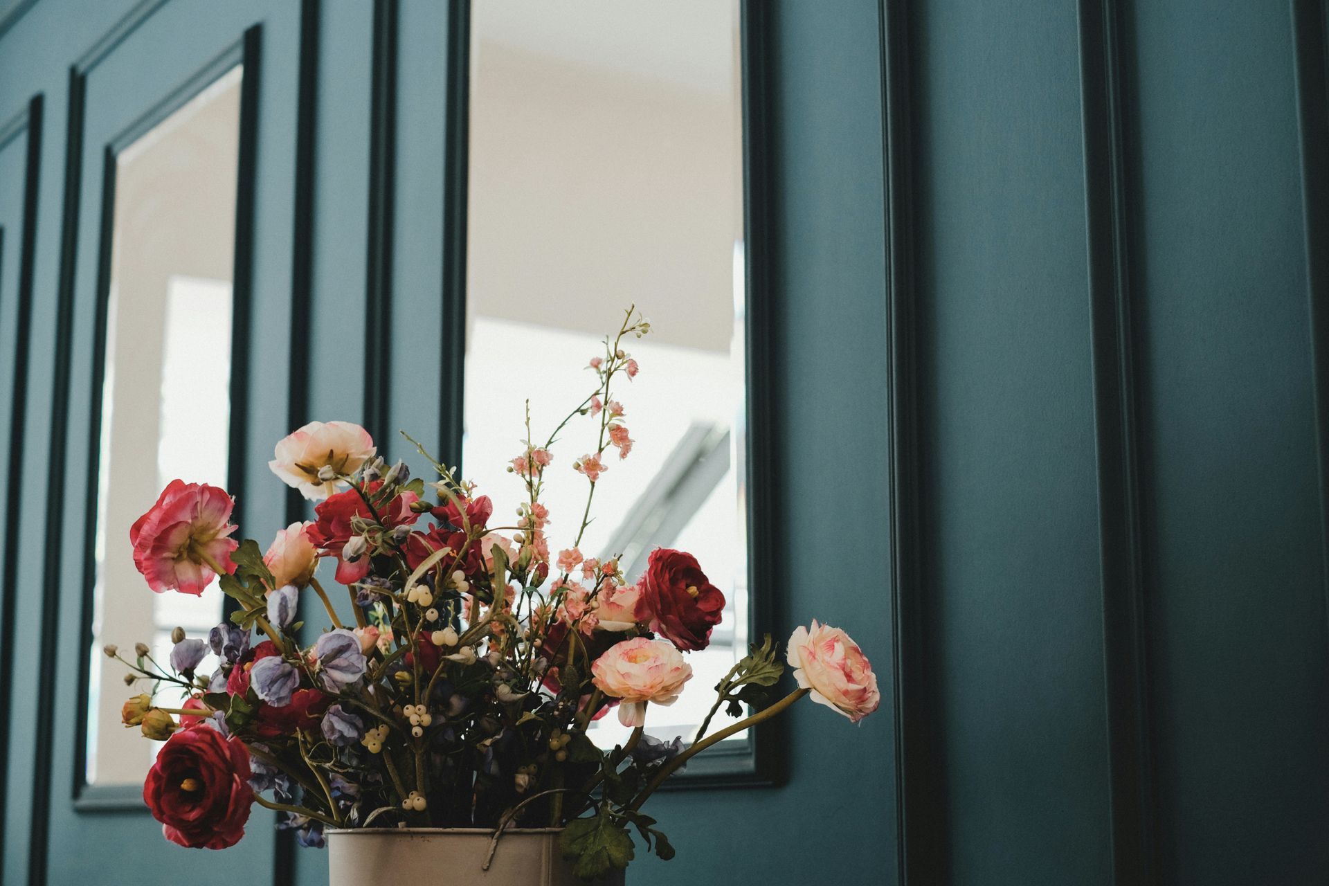 Bouquet of colorful flowers in vase in front of a teal paneled wall with a mirror.