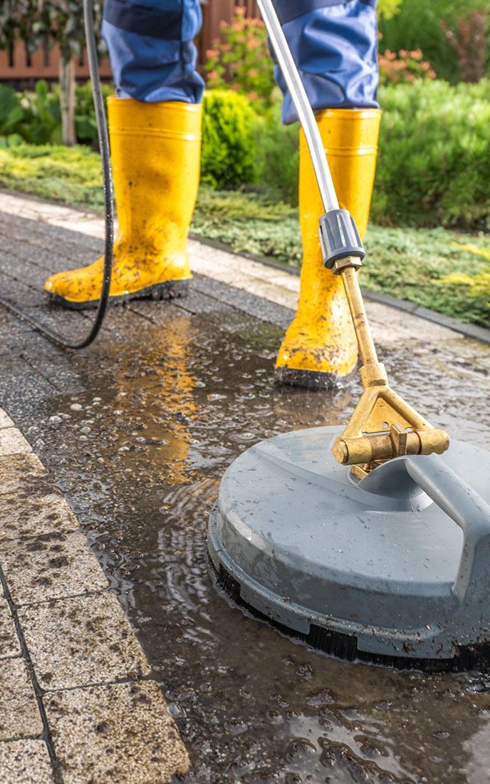 Person in yellow boots power washing a dark brick patio.