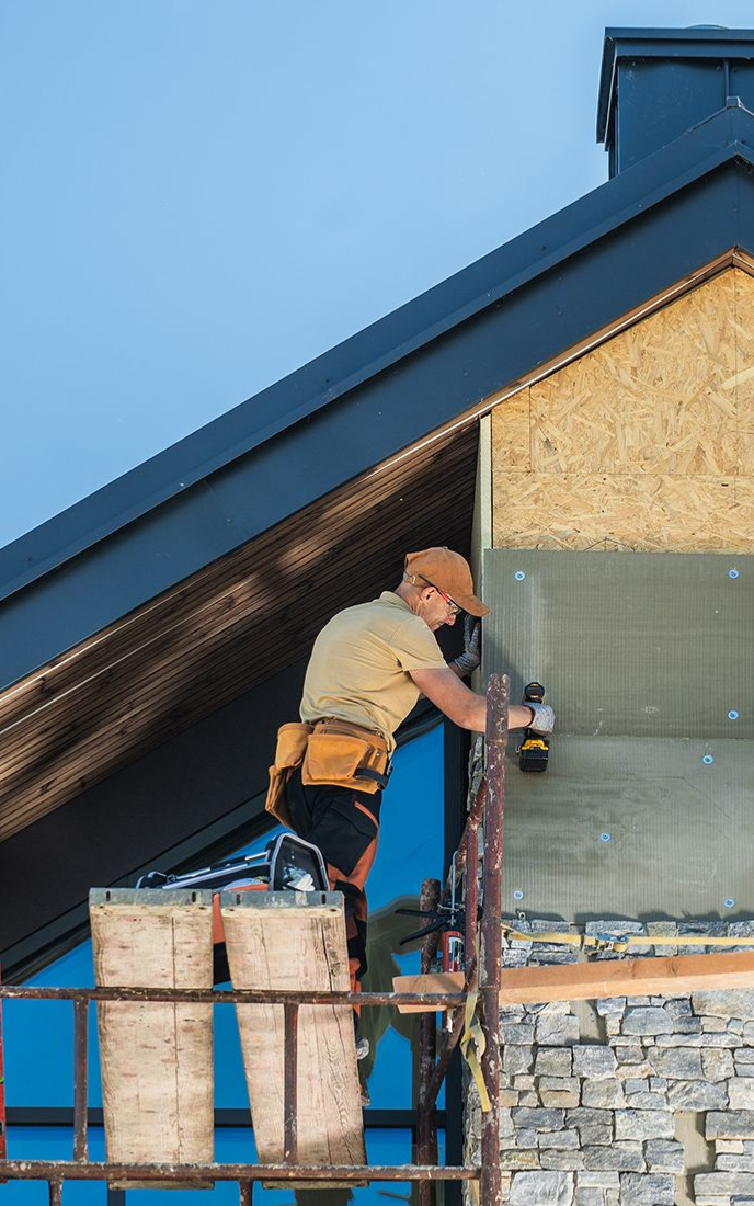 Construction worker on scaffolding installing insulation on a building under a blue sky.