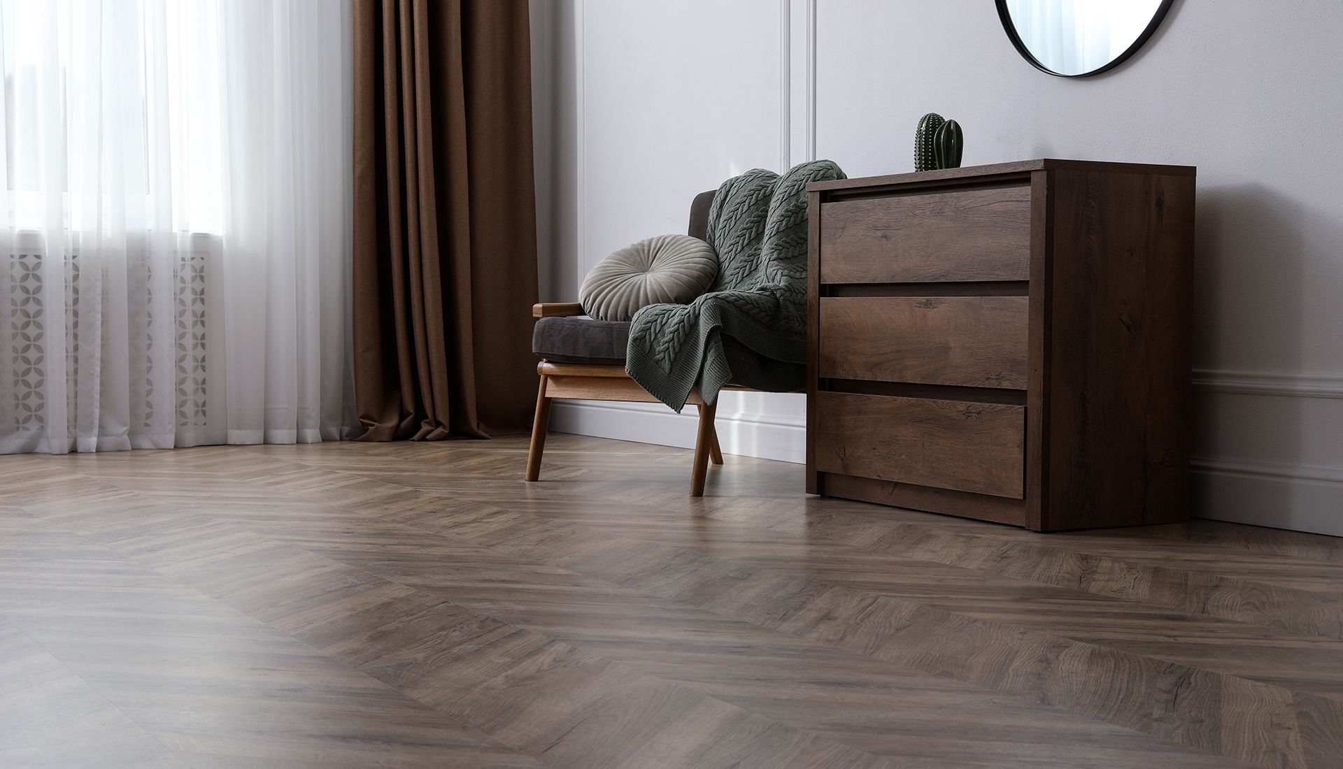 Wooden chest of drawers next to a chair with a blanket and pillow. Room with hardwood floor and window.