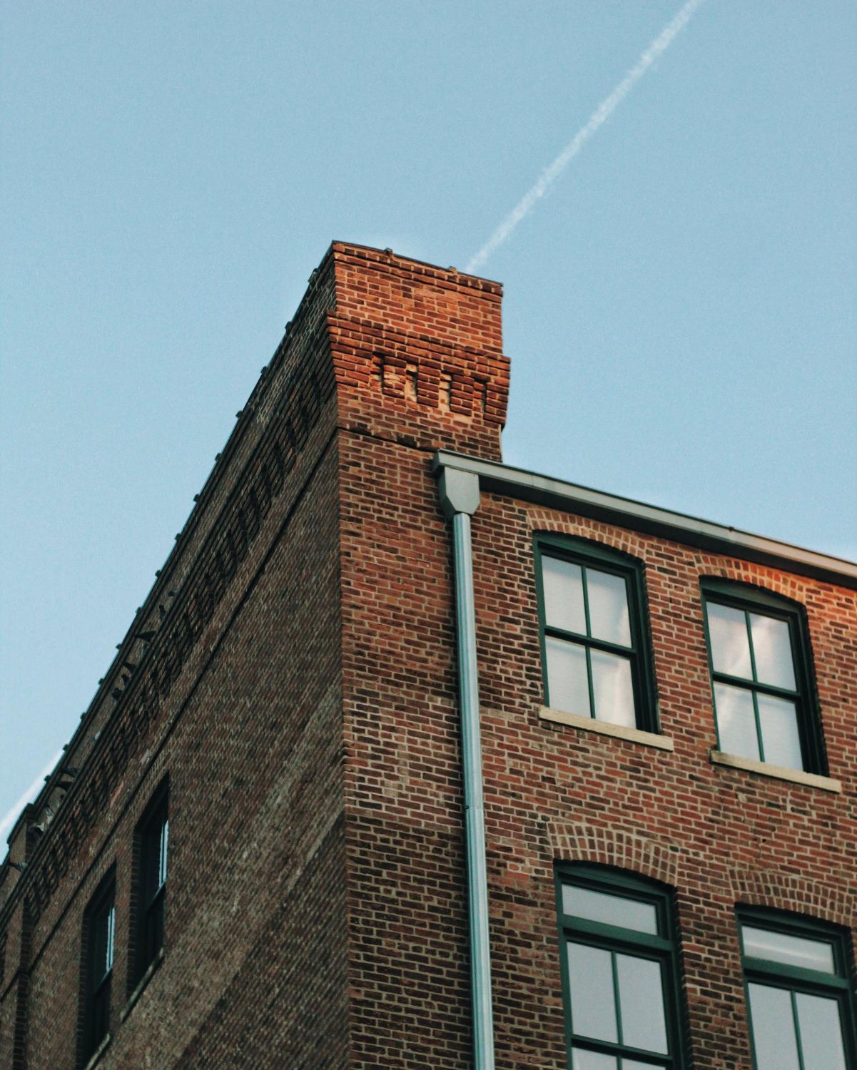 A low-angle view of a brick building corner featuring a decorative cornice and a vertical metal drainpipe against a blue sky.
