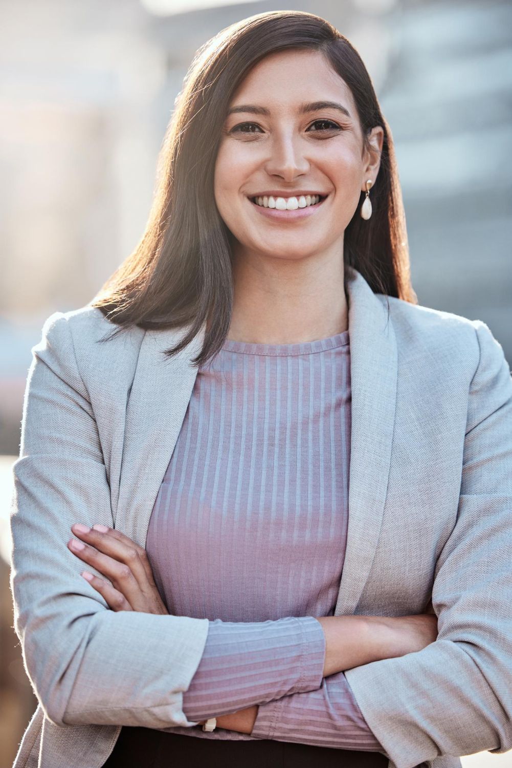A smiling woman with long dark hair, wearing a mauve top and light blazer with arms crossed, stands in an office setting.
