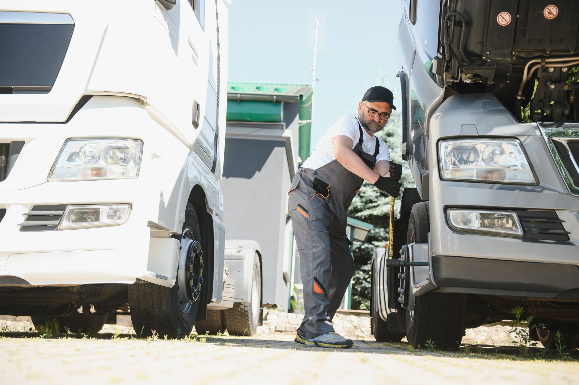 A mechanic wearing grey work overalls inspects the engine of a dark truck parked next to a white truck outdoors.