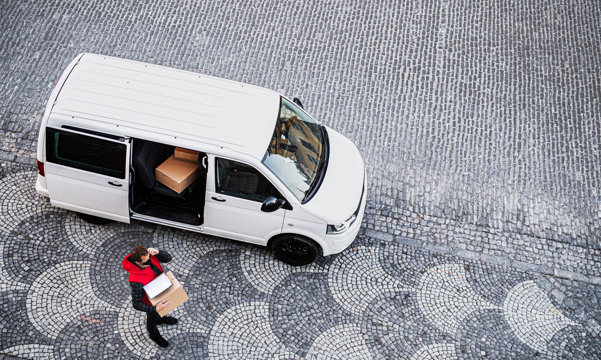 A delivery person in a red jacket carries boxes from a white van parked on a cobblestone street, viewed from above.