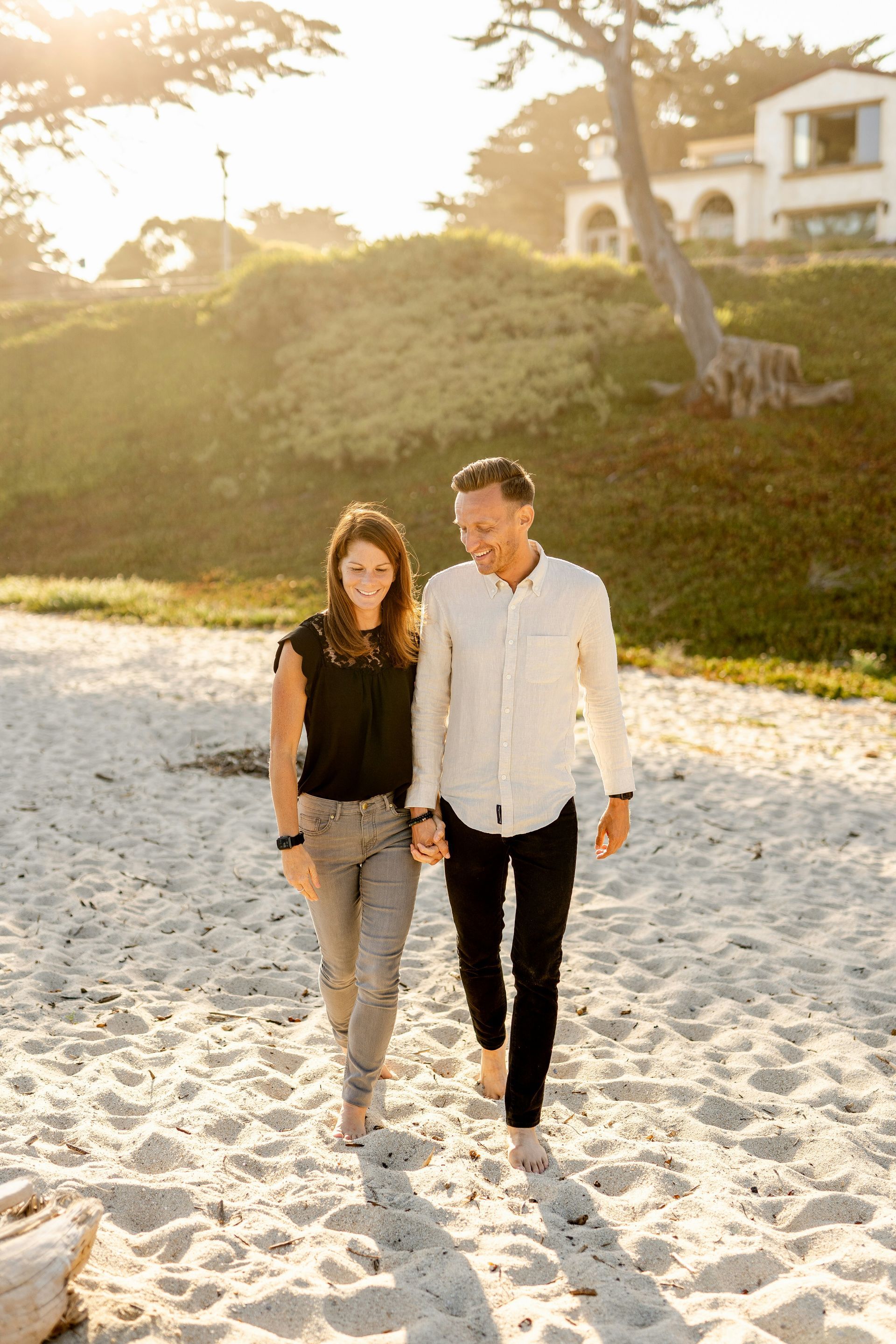 A couple walks hand-in-hand across a sandy beach at sunset, with a grassy hill and a building in the background.