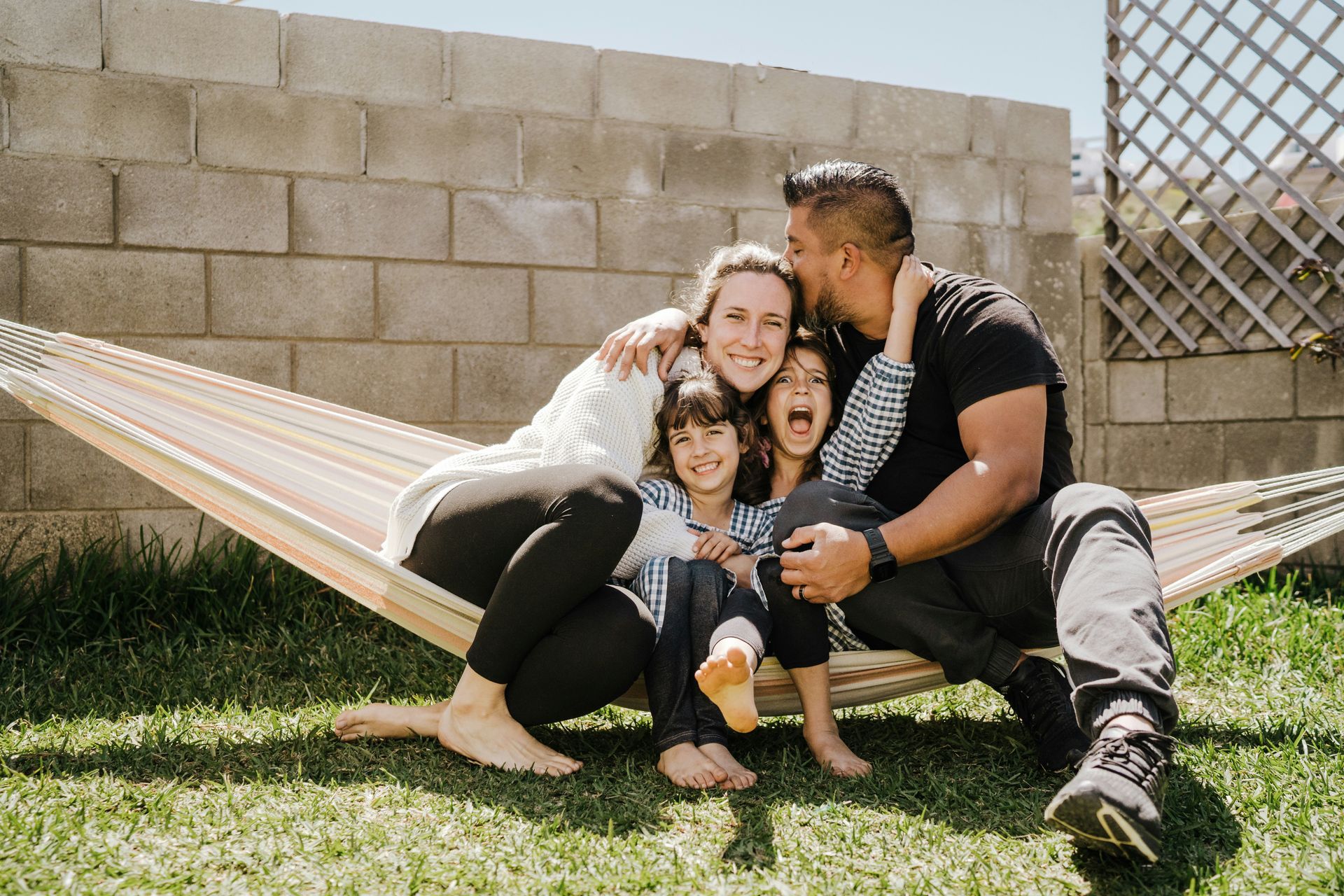 A family sits together in a hammock in a sunny backyard, laughing and embracing in front of a block wall.