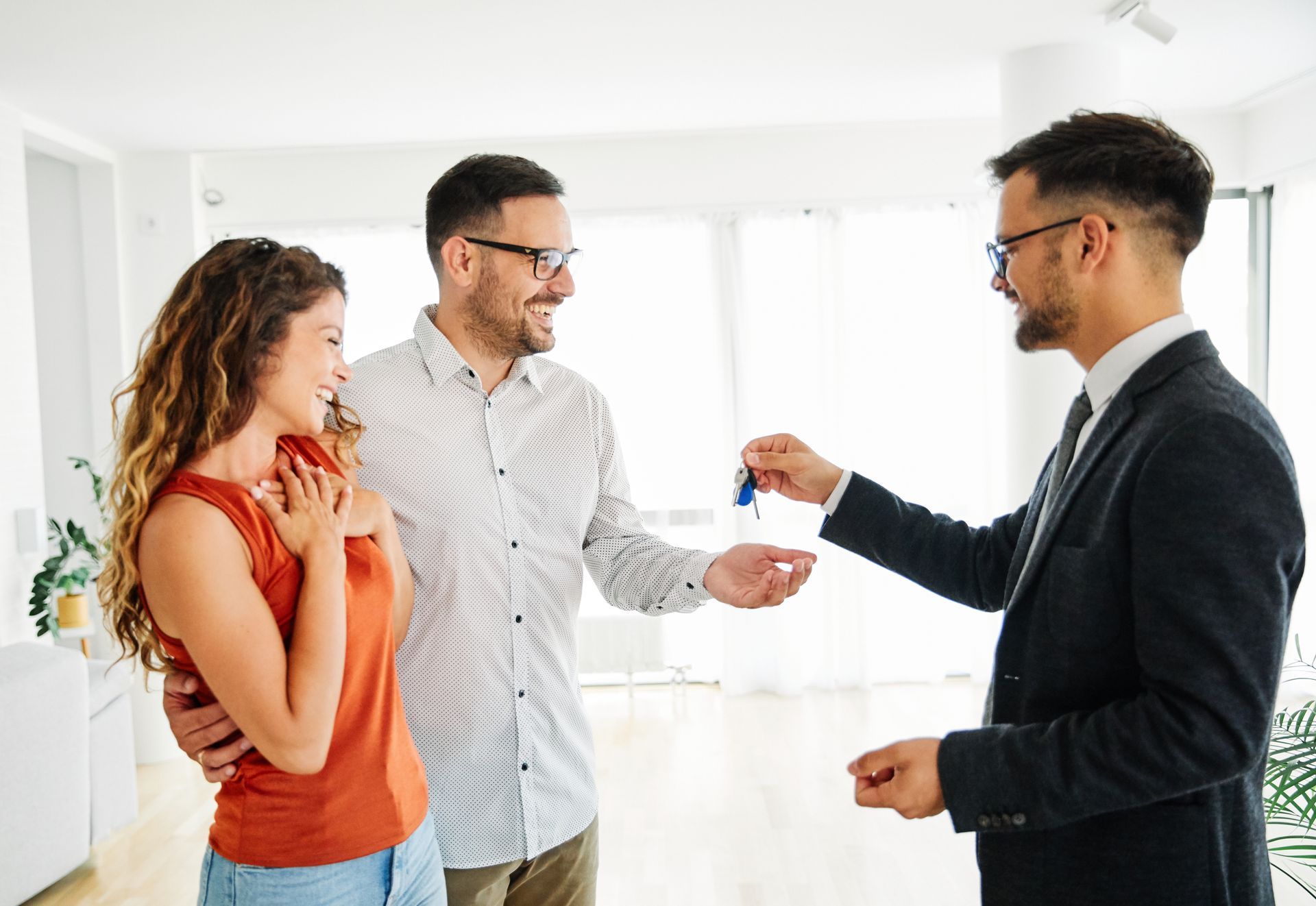 A real estate agent hands keys to a smiling couple in a bright, modern, unfurnished room.