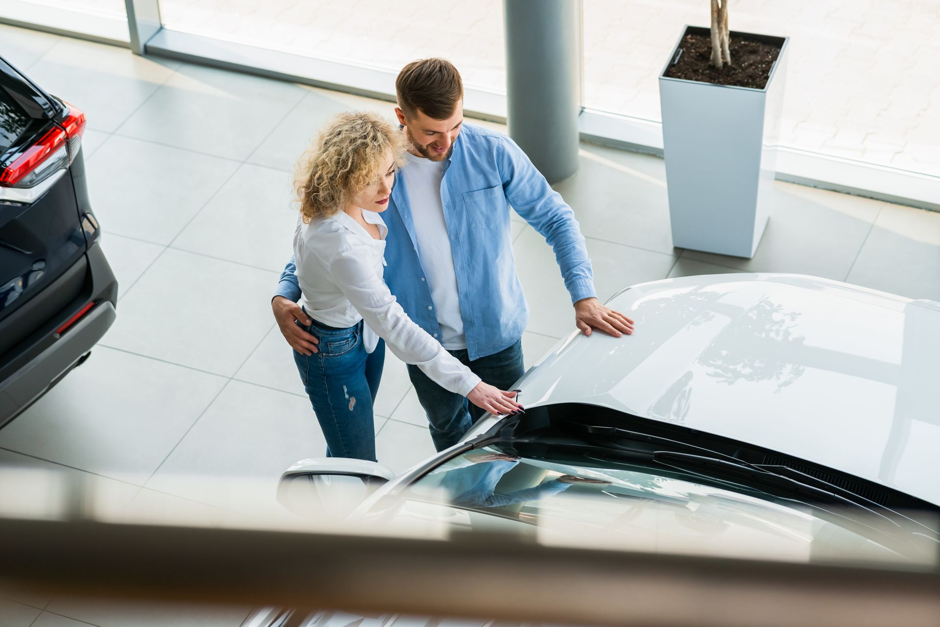 Two people in a dealership showroom leaning over the hood of a white car, looking at the vehicle.