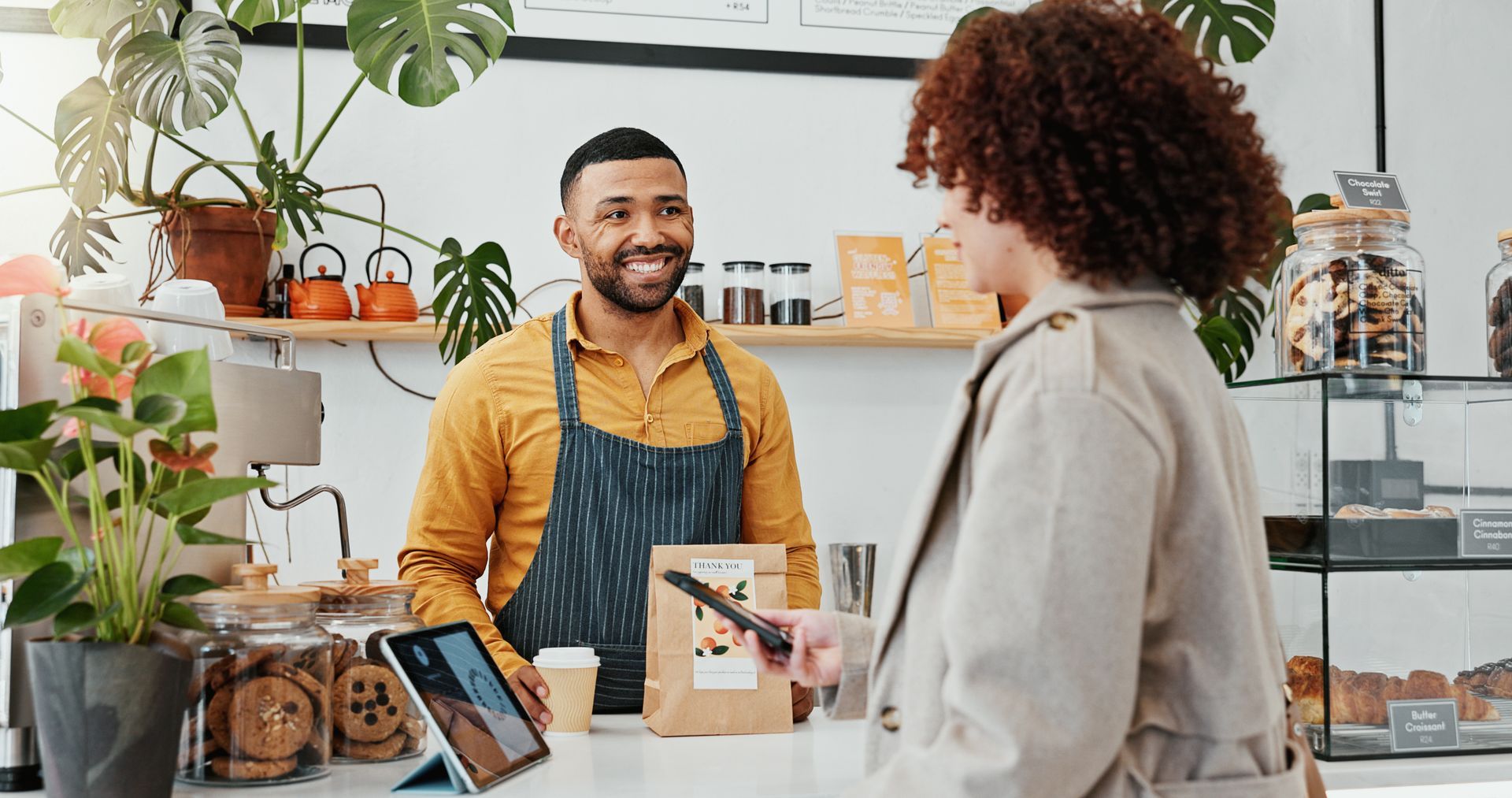 A smiling barista in an apron helps a customer at a cafe counter while she holds a card terminal for payment.