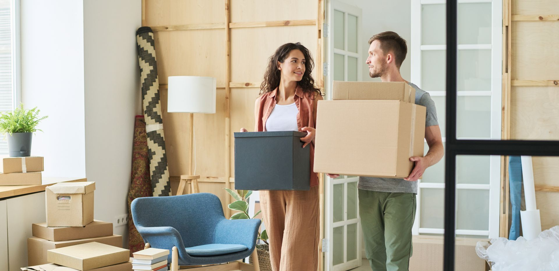 A couple moves into a new home, carrying cardboard boxes in a bright, sparsely furnished room with a blue armchair.
