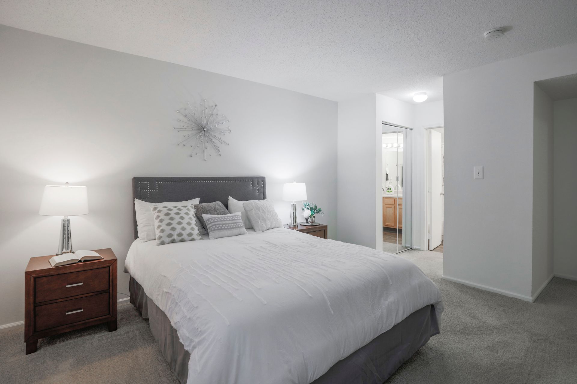 Bright bedroom with a white bed, gray carpet, bedside lamps, and a bathroom visible through an open doorway